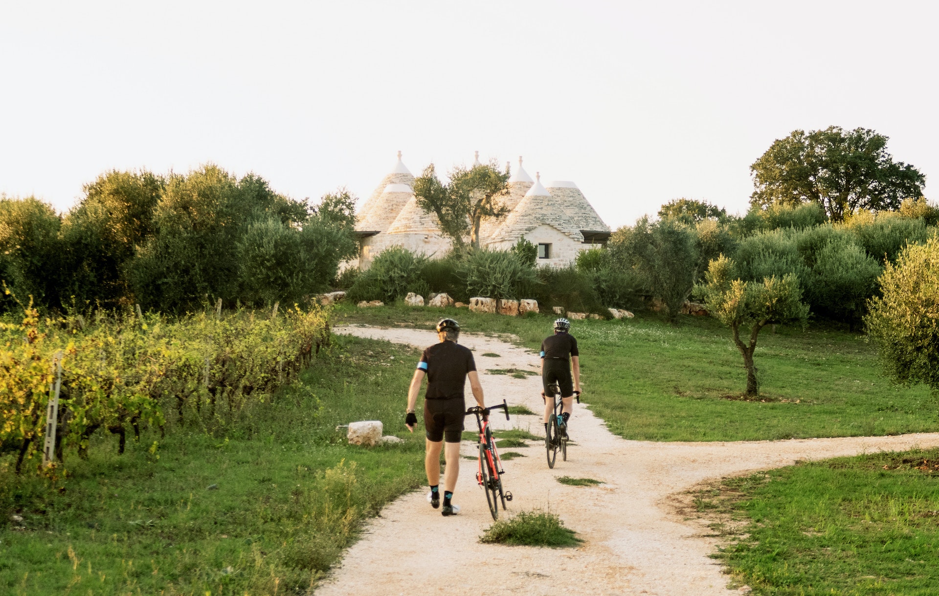 Two cyclists walk their bikes down a path towards some conical white limestone buildings