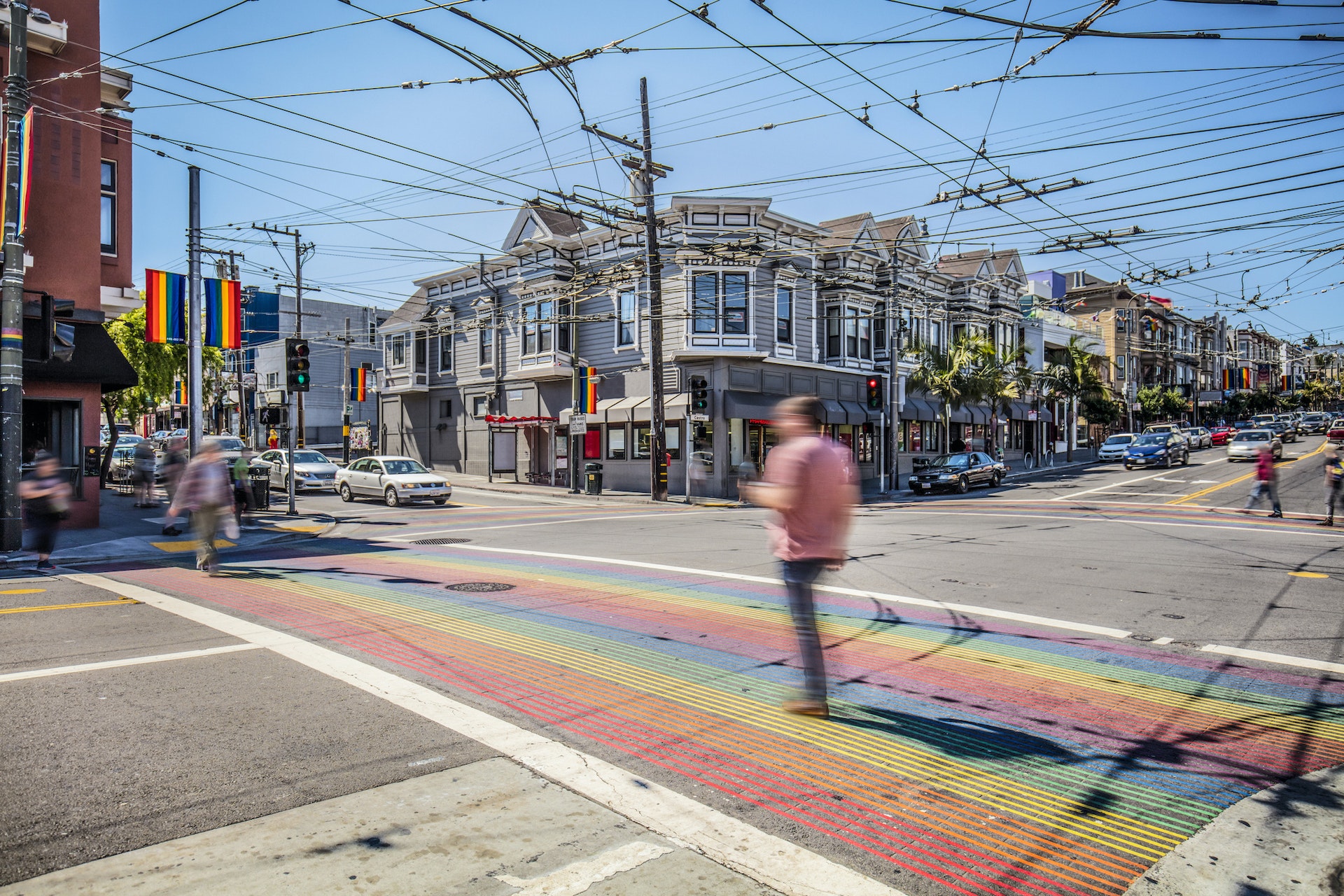 People cross the road on a rainbow-colored crosswalk in a city neighborhood. Rainbow flags fly from buildings