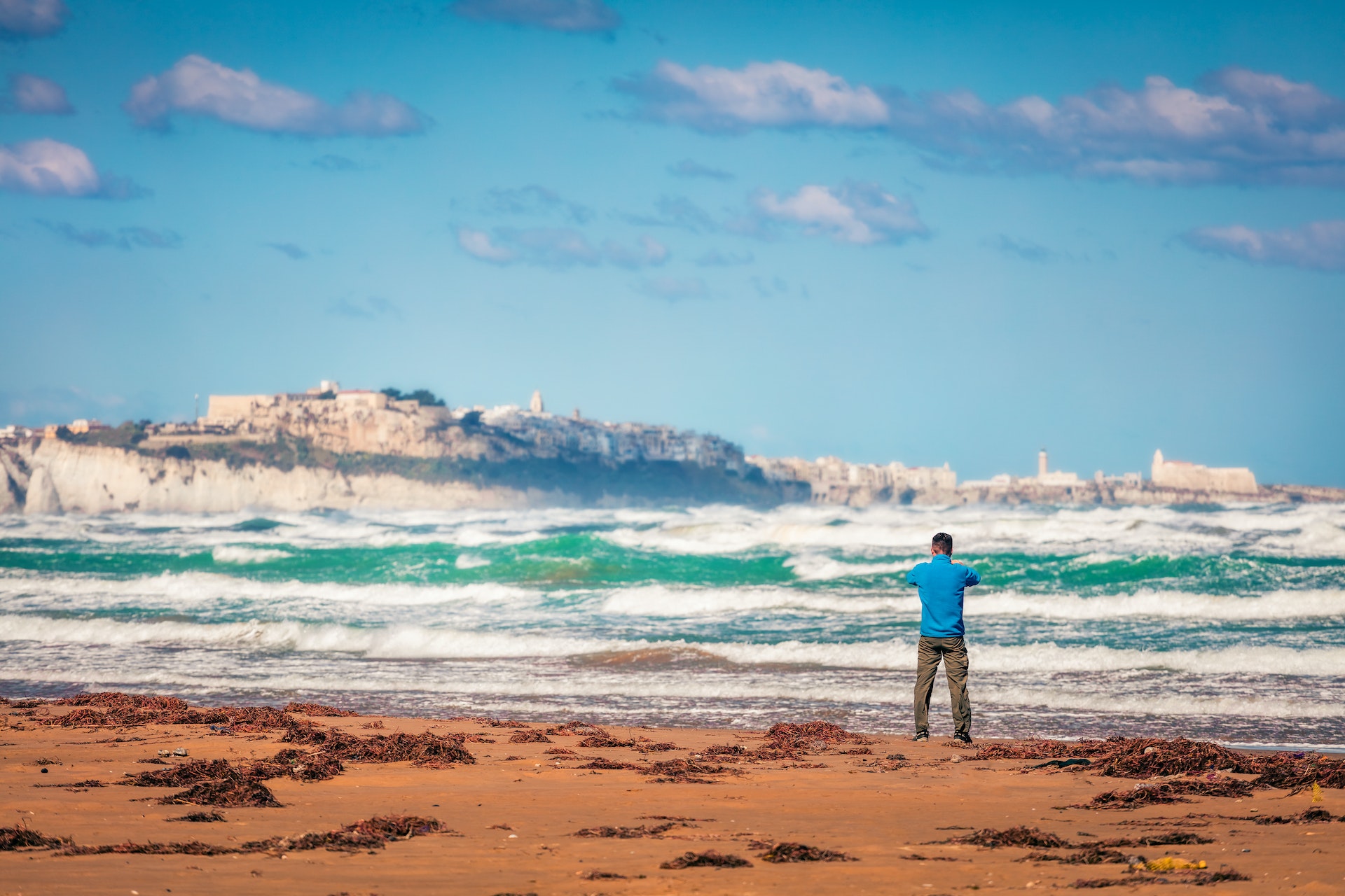 A man stands on a beach with waves crashing at sea and takes a photo of a clifftop town in the distance