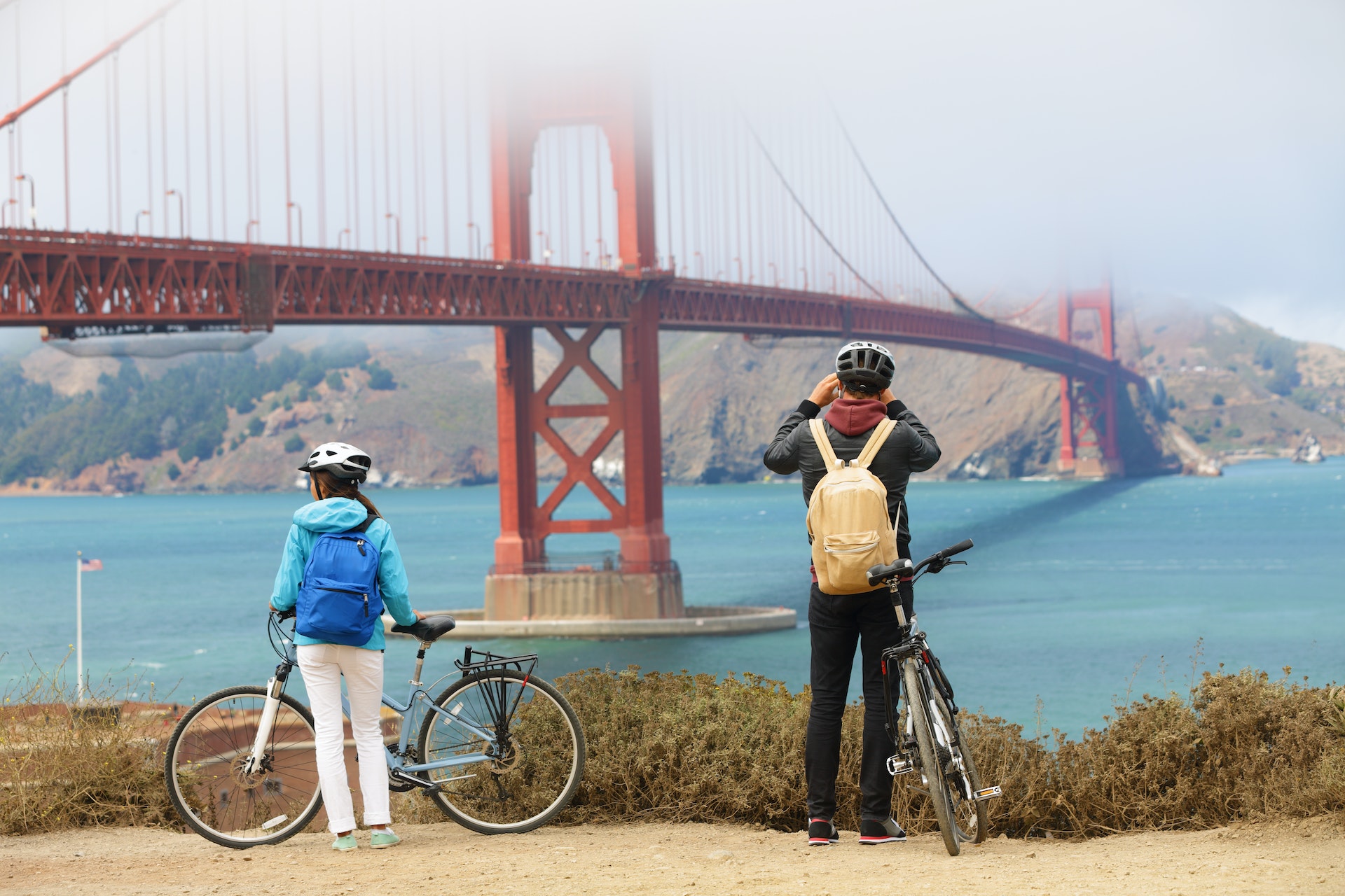A couple with bikes pause on a trail to take photos of a large orange-red bridge shrouded in fog