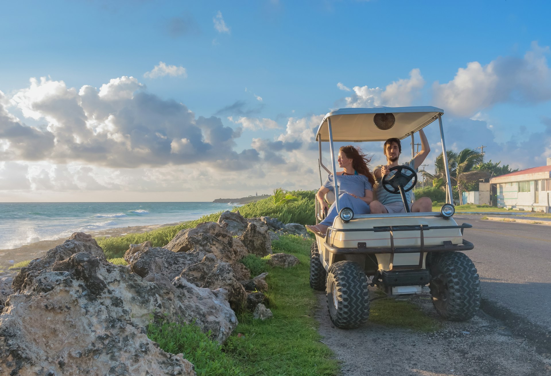 A young couple driving a golf cart alongside a tropical beach 