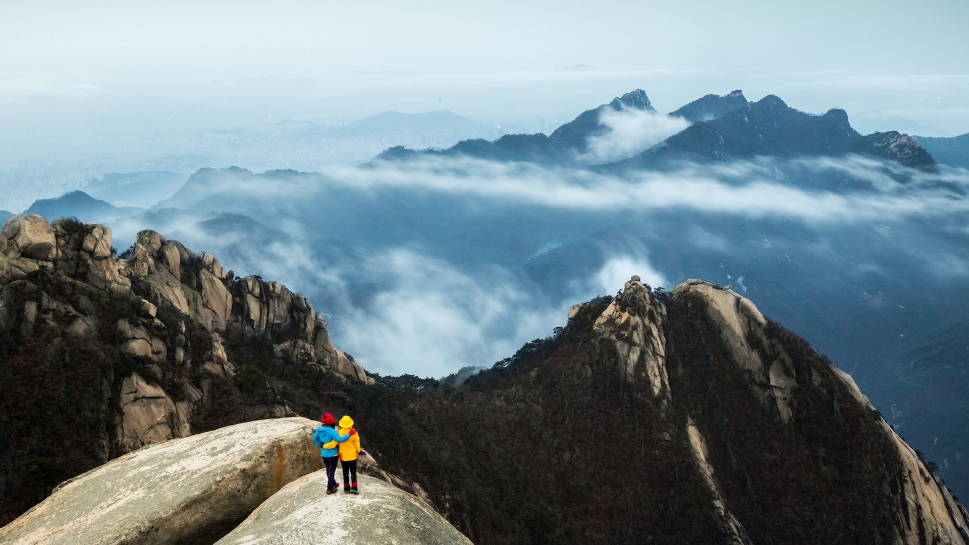 An aerial view of two hikers on a rocky outcrop overlooking cloudy mountain peaks a