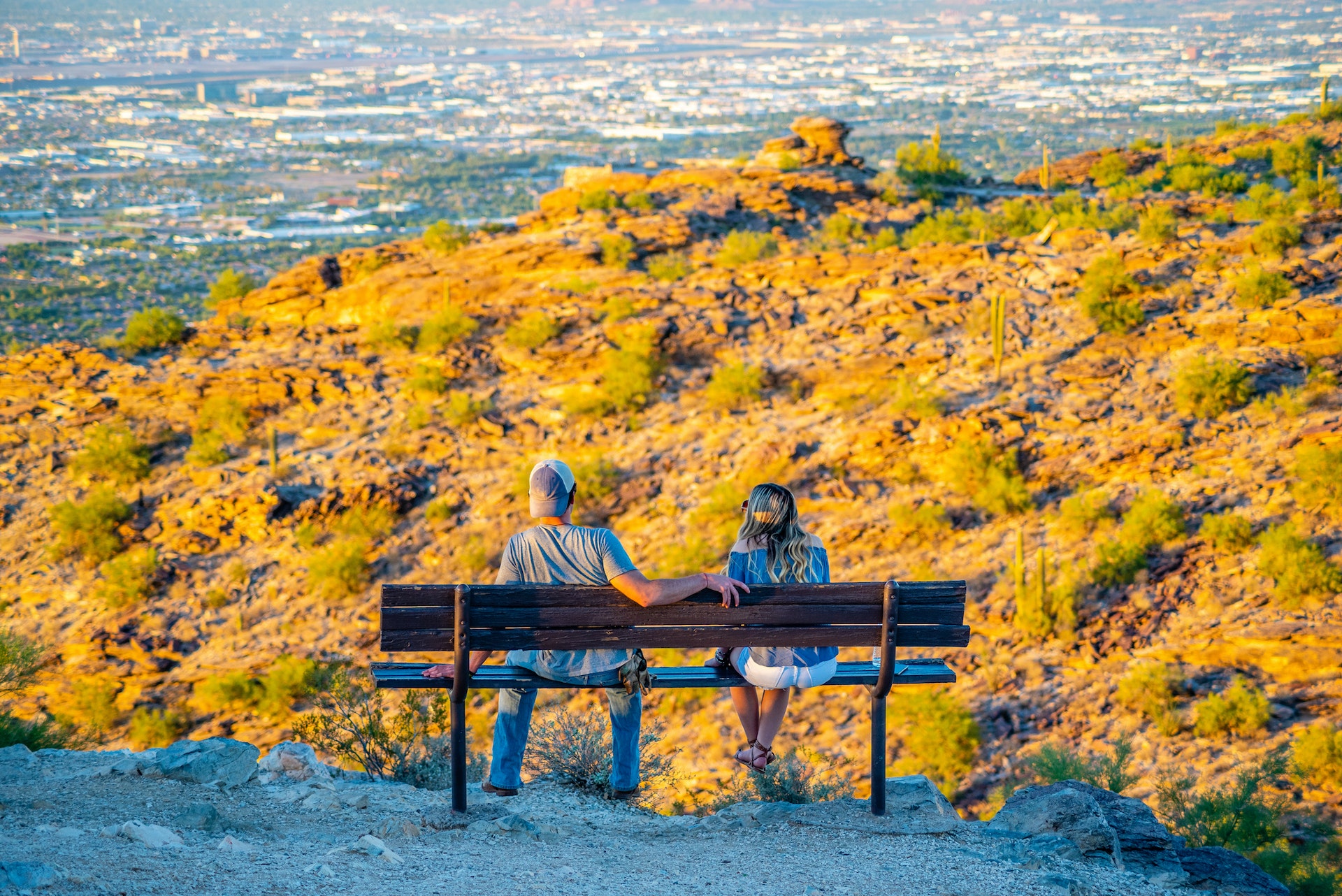 A couple sit on a bench at a lookout with the sun shining down over an arid landscape